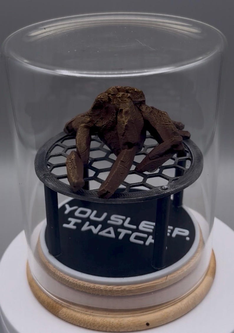 May include: A brown, textured, crab-like figurine sits on a black, circular, honeycomb-patterned platform inside a clear glass dome. The base features the white text "YOU SLEEP. I WATCH." The entire display rests on a light-colored wooden circular stand.