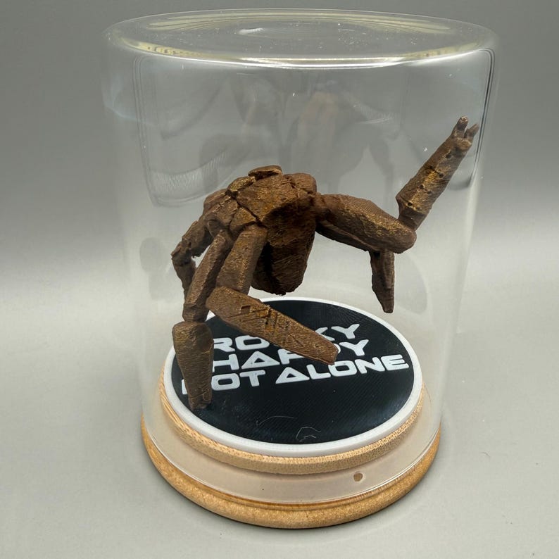 May include: A brown, textured, multi-legged creature figurine enclosed in a clear glass dome on a light wood base. The circular black display stand inside features the white text "ROCKY HARDY NOT ALONE" printed in a bold, sans-serif font.