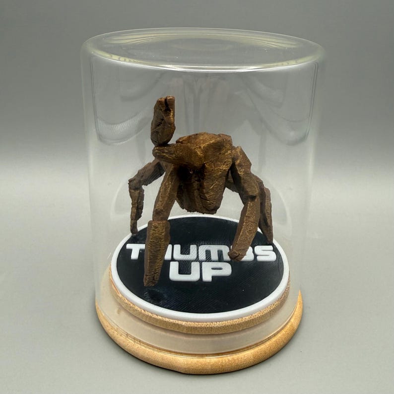 May include: A bronze-colored, multi-legged creature figurine with a textured surface, positioned inside a clear glass display dome. The figure rests on a circular black base featuring the white text THUMBS UP, set upon a light wooden stand.