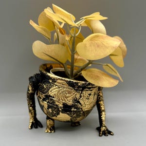 May include: A decorative planter with a black and gold finish, featuring a unique design with legs. The planter holds a faux plant with yellow-green leaves, adding a touch of nature to the artistic piece. The pot has a textured surface.