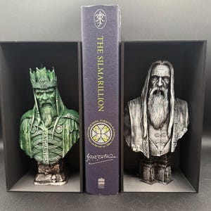 May include: A set of bookends featuring busts of characters from The Lord of the Rings, flanking a copy of "The Silmarillion." The bookends are in black boxes, with one bust in green and the other in silver. The book's spine is dark blue.