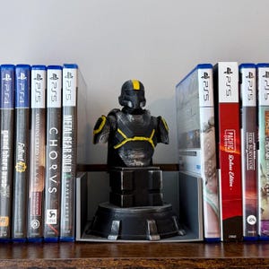 Puede incluir: Un busto negro y amarillo de un soldado futurista se encuentra en una estantería, flanqueado por cajas de videojuegos. El busto tiene un diseño detallado con detalles amarillos. Los videojuegos incluyen "Metal Gear Solid" y "Jedi Survivor".
