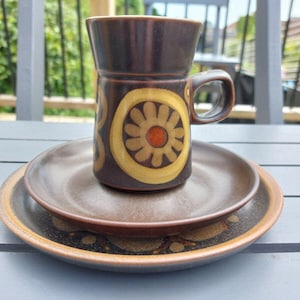 May include: A brown ceramic coffee mug with a yellow and orange floral design sits on a brown saucer. The saucer is on top of a larger brown saucer with a similar design.