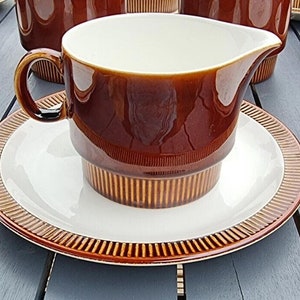 Puede incluir: Una salsera de cerámica marrón y blanca con un platillo. La salsera tiene un diseño acanalado y un interior blanco. El platillo tiene un borde marrón con una franja dorada.