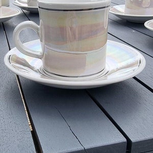 May include: A set of white teacups and saucers with a pearlescent iridescent glaze. The cups and saucers have a black band around the rim. The cups and saucers are on a gray wooden table.