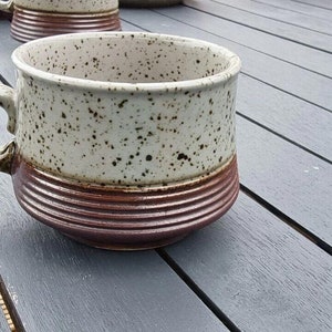 Könnte beinhalten: Eine gesprenkelte braune und weiße Keramiktasse mit einem braunen Henkel. Die Tasse hat einen braunen Streifen um den oberen Rand und einen braunen Streifen um den unteren Rand.