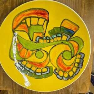 Può includere: Un piatto rotondo in ceramica con uno sfondo giallo brillante caratterizzato da un design astratto centrale nelle tonalità dell'arancione, del verde lime e del blu scuro. Il motivo grafico è composto da linee curve e forme geometriche.
