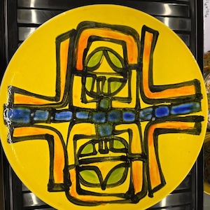 Può includere: Un piatto rotondo in ceramica con uno sfondo giallo brillante caratterizzato da un audace design astratto in nero, arancione e blu. Il motivo centrale è composto da forme geometriche e linee, tra cui una fascia blu orizzontale e accenti verdi.