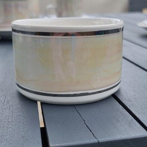 May include: A white ceramic bowl with a pearlescent, iridescent finish. The bowl has a black stripe at the top and bottom. The bowl is sitting on a dark gray wooden table. Other dishware is visible in the background.
