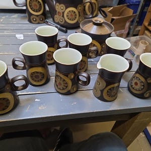 May include: A vintage brown ceramic tea set with a floral design. The set includes mugs, a creamer, a teapot, and a sugar bowl. The mugs have a dark brown exterior with a cream-coloured interior and a decorative yellow and brown floral pattern.