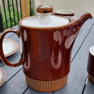 May include: A brown ceramic teapot with a white lid and a gold band around the bottom. The teapot has a rounded body and a spout on the side.