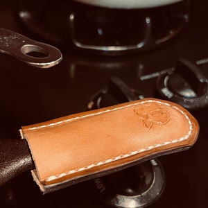 May include: A tan leather handle cover with white stitching and a stamped design. The cover is on a black pan handle, with a white pot in the background. The handle cover is designed to protect hands from heat.