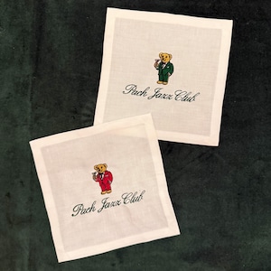Puede incluir: Dos posavasos cuadrados de lino blanco con diseños bordados. Cada posavasos presenta un oso de dibujos animados con traje sosteniendo una bebida, con el texto "Pach Jazz Club" en verde. Un oso lleva un traje verde, el otro un traje rojo.