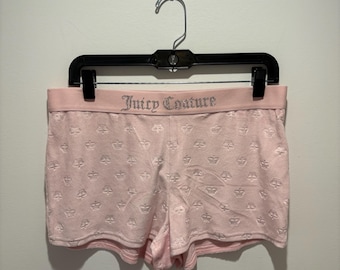 May include: Pink shorts with a repeating crown pattern and a pink waistband. The waistband features the text "Juicy Couture" in silver lettering. The shorts are hanging on a black hanger against a white wall.