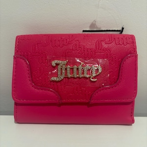 May include: A vibrant pink wallet featuring the word "Juicy" in gold and rhinestone lettering. The wallet has a flap closure and a textured pattern with the word "Juicy" embossed throughout. A small tag hangs from the top.