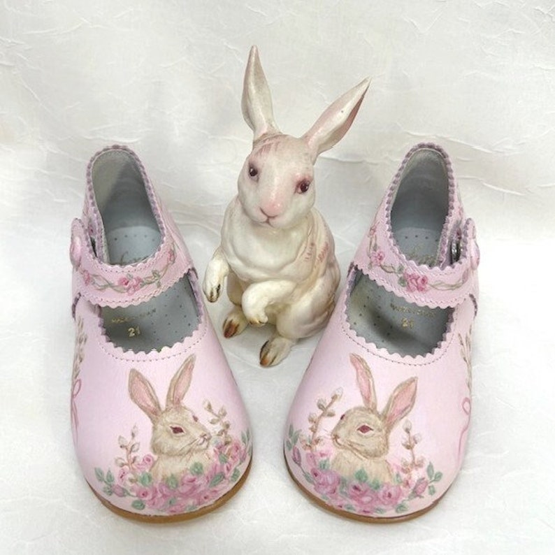 Toes are adorned in a  composition of a bunny, pussy willows, and roses. Tan and brown bunnies on each toe face each other. The entire top of the shoe and the strap going across the instep are scalloped and have a pink garland. Background is pink.