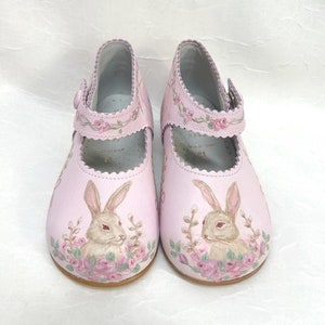 Toes. Pink background.  One brown and tan bunny on each toe and face each other.  Bunnies are nestled in pink roses, pussy willows, and greenery. The top edge and strap are scalloped.  A rose garland runs across the strap and around the top of shoe.