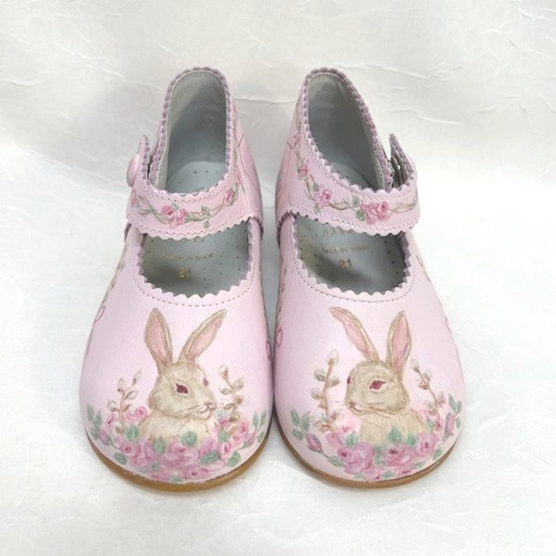 Toes. Pink background.  One brown and tan bunny on each toe and face each other.  Bunnies are nestled in pink roses, pussy willows, and greenery. The top edge and strap are scalloped.  A rose garland runs across the strap and around the top of shoe.