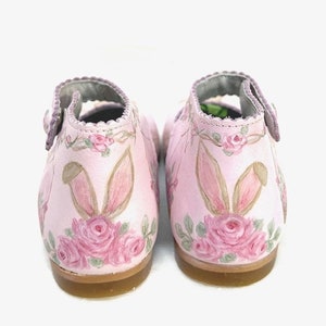 The back sports bunny ears peeking up from a spray of roses and foliage.  You can see a rose garland running around the top of the scalloped edge.  The background is soft pink and the bunny ears are brown and tan with shades of pink inside the ears.