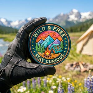 Op de afbeelding: Een ronde patch met de woorden "WILD & FREE" en "STAY CURIOUS" rond een berg- en kampvuurscène. De patch wordt getoond tegen een achtergrond van bergen, een tent en een rivier.