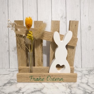 Könnte beinhalten: Osterdekoration aus Holz mit einer weißen Hasen-Silhouette und einem Herz-Ausschnitt, vor einem rustikalen Holzzaun. Eine kleine Glasvase enthält getrocknete gelbe Blumen. Der Schriftzug "Frohe Ostern" ist grün.