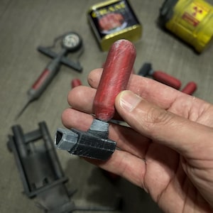 May include: A red and black device with a cylindrical red component and a black rectangular base. Other items in the image include a syringe, a yellow container with a radiation symbol, and a can of food.