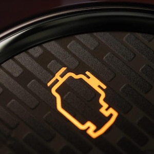 Può includere: Primo piano del cruscotto di un'auto con il simbolo arancione illuminato della spia del motore. Il simbolo è un contorno stilizzato di un motore, che indica un potenziale problema meccanico. Lo sfondo è una superficie strutturata grigio scuro.