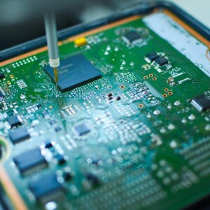 May include: Close-up of a green circuit board with various electronic components. A tool is positioned over a chip, possibly for testing or repair. The board has a complex layout with multiple integrated circuits and connectors.