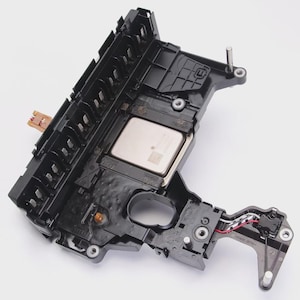 May include: A black automotive transmission control module with various components, including a silver-colored processor and multiple connectors. The module has a complex, multi-layered design with several mounting points and wiring.