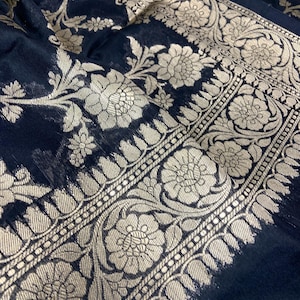 May include: Close-up of a dark navy fabric with intricate floral patterns in a metallic gold color. The design features large flowers and detailed borders, creating a luxurious textile appearance.