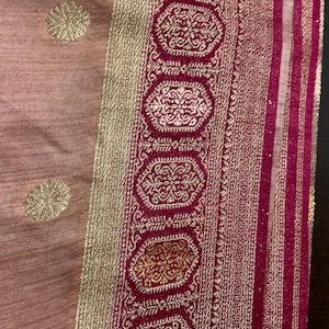 May include: A close-up of a pink fabric with a gold and red border. The border has a repeating pattern of small circles and a larger circle with a floral design. The fabric is likely a sari or other traditional Indian garment.