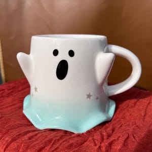 May include: White ceramic ghost mug with a black face and a blue ombre bottom. The mug has silver stars on the side.