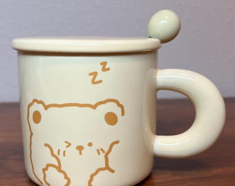 May include: A cream-colored ceramic mug with a brown bear illustration and the word "zz" on the side. The mug has a lid and a spoon.
