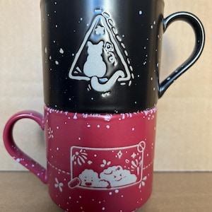 May include: Two stacked ceramic mugs. The top mug is black with white speckles and a cat design inside a triangle. The bottom mug is red with white speckles and a cloud design. Both mugs have handles.