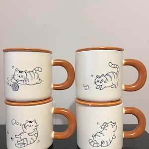 May include: Set of four ceramic mugs with brown handles. Each mug features a different illustration of a cat with a white background. The mugs are stacked in two pairs.
