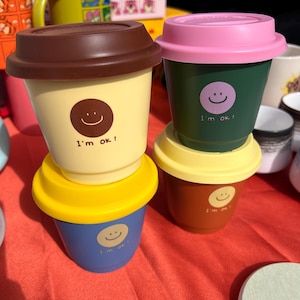 May include: Four reusable coffee cups with lids. Each cup has a different color and a smiley face with the text "I'm ok!" printed on it. The cups are stacked on top of each other.