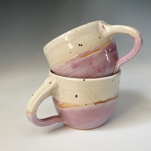 Two wheel-thrown pottery cups with cozy round shapes, smooth rims, and shapely slim handles. Lightweight, nice feel in hand. Hold eight ounces each. Pink and white colors.