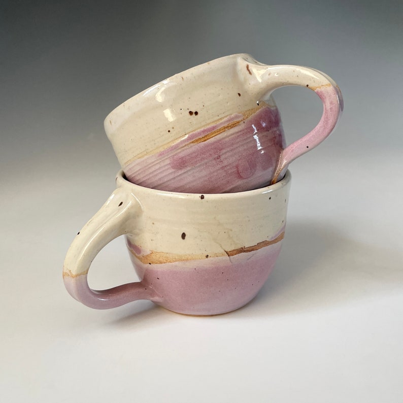 Two wheel-thrown pottery cups with cozy round shapes, smooth rims, and shapely slim handles. Lightweight, nice feel in hand. Hold eight ounces each. Pink and white colors.