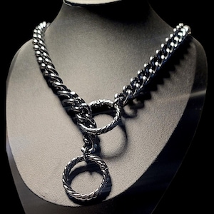 May include: A dark metallic chain necklace featuring a thick, interlocking curb link design. The necklace includes a central circular ring connector that holds a secondary, smaller twisted circular pendant hanging from the main chain structure.