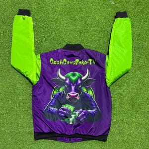 May include: A purple and lime green bomber jacket featuring a graphic on the back of a bull with green hair holding stacks of cash. The text CashCowsPokerTV is printed in lime green above the bull illustration. The jacket has long lime green sleeves.