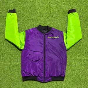 May include: A purple bomber jacket with bright lime green sleeves, a black ribbed collar, black cuffs, and a black front zipper. The text CashCowsPokerTV is embroidered in lime green on the upper left chest area of the jacket's purple front panel.