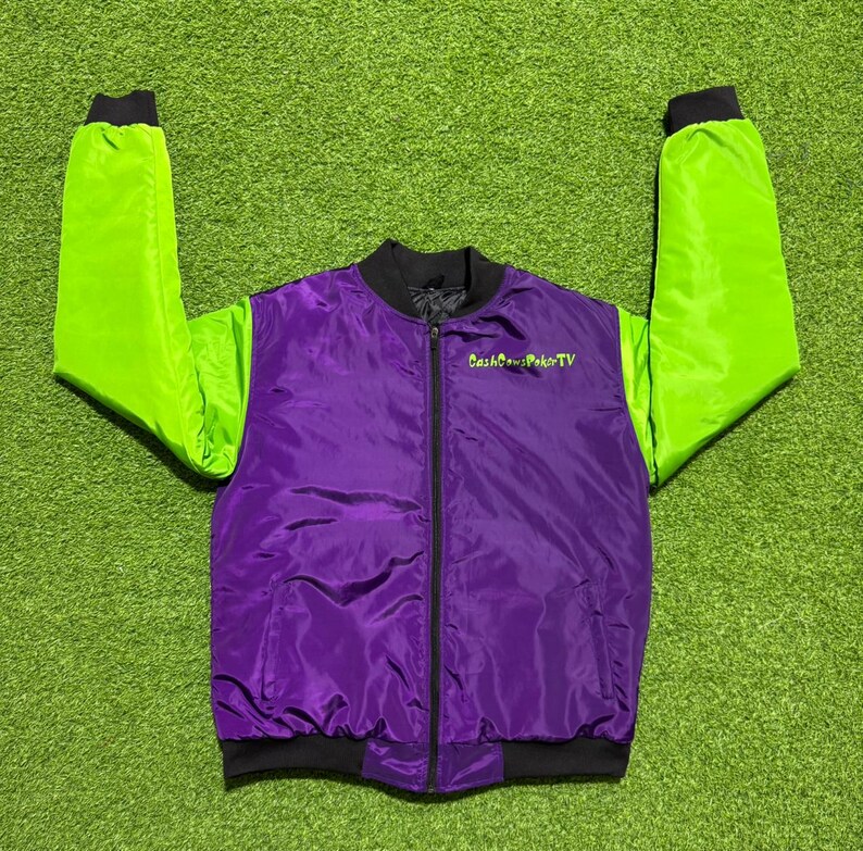 May include: A purple bomber jacket with bright lime green sleeves, a black ribbed collar, black cuffs, and a black front zipper. The text CashCowsPokerTV is embroidered in lime green on the upper left chest area of the jacket's purple front panel.