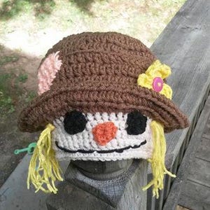 May include: A crocheted scarecrow hat with a brown brim, white face, black eyes, orange nose, and yellow yarn hair. The hat has a pink and yellow flower on the side.