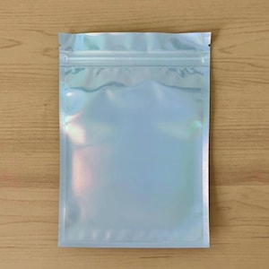 Puede incluir: Una bolsa transparente, iridiscente y resellable con cierre de cremallera. La bolsa tiene forma rectangular y est&aacute; vac&iacute;a. La superficie refleja colores como el azul, el verde y el morado. La bolsa est&aacute; sobre una superficie de madera marr&oacute;n claro.