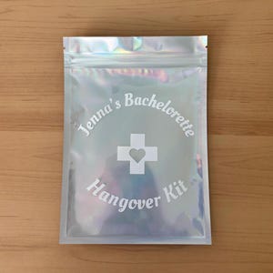 Puede incluir: Bolsa iridiscente con cremallera con el texto "Jenna's Bachelorette Hangover Kit" en escritura blanca. Una cruz blanca con un coraz&oacute;n en el centro est&aacute; centrada en la bolsa. La bolsa est&aacute; sobre una superficie de madera marr&oacute;n claro.