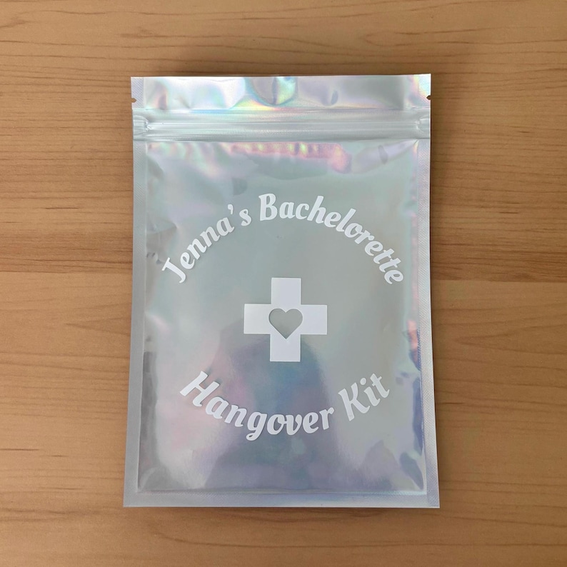 Puede incluir: Bolsa iridiscente con cremallera con el texto "Jenna's Bachelorette Hangover Kit" en escritura blanca. Una cruz blanca con un coraz&oacute;n en el centro est&aacute; centrada en la bolsa. La bolsa est&aacute; sobre una superficie de madera marr&oacute;n claro.