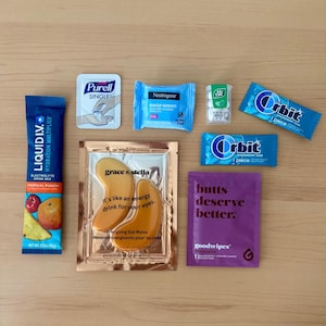 Puede incluir: Surtido de art&iacute;culos de cuidado personal sobre una superficie de madera. Incluye un sobre de mezcla para bebidas Liquid I.V. azul, desinfectante de manos Purell, toallitas desmaquillantes Neutrogena, caramelos de menta Tic Tac, chicle Orbit, mascarillas para ojos y goodwipes.