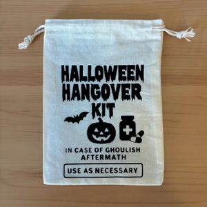 Puede incluir: Una bolsa de algodón color crema con cordón y texto en negro que dice HALLOWEEN HANGOVER KIT, IN CASE OF GHOULISH AFTERMATH y USE AS NECESSARY. El diseño incluye una silueta negra de un murciélago, una calabaza y un frasco de medicina.