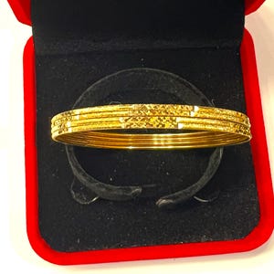 Puede incluir: Un conjunto de brazaletes finos de oro con un patrón texturizado de corte de diamante en la superficie. Las pulseras están apiladas, mostrando un acabado metálico reflectante y una forma circular consistente, presentadas sobre un inserto de terciopelo negro.