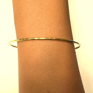 Pode incluir: Uma pulseira tipo bracelete circular e fina em ouro, com uma superfície martelada e texturizada que reflete a luz. A banda de metal tem um acabamento polido e um perfil fino e consistente, criando uma aparência simples e brilhante.